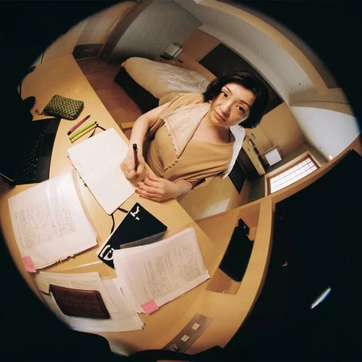 A fish-lens photograph of author Murata Sayaka with papers and other writing utensils sprawled around her.
